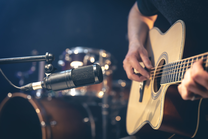 Male musician playing acoustic guitar behind microphone in recording studio.