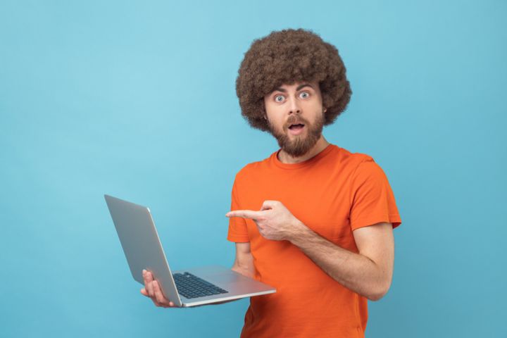Man pointing laptop screen and looking at camera with shocked expression, open mouth.
