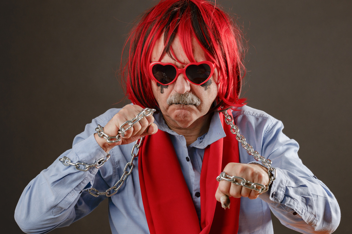 An elderly man with red hair wearing a red scarf and heart-shaped glasses stands in a scary stance with a metal chain