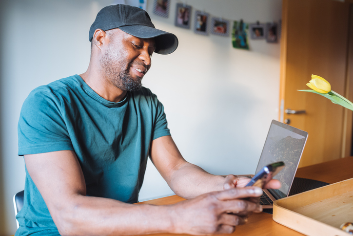 Contented black man sitting at table where standing open laptop, holding smartphone and looking at screen with relaxed smile and friendly expression.