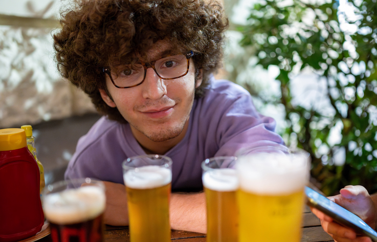 Portrait of a smiling young Caucasian man with curly hair and glasses: the beers have just been brought to the table, the evening with friends begins. Friendship, conviviality, joy of life.