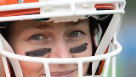 A woman is geared up in a football helmet, displaying a confident smile. She has black eye paint and is ready for the upcoming game, embodying passion for the sport