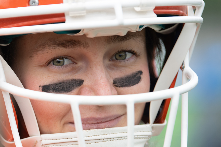 A woman is geared up in a football helmet, displaying a confident smile. She has black eye paint and is ready for the upcoming game, embodying passion for the sport