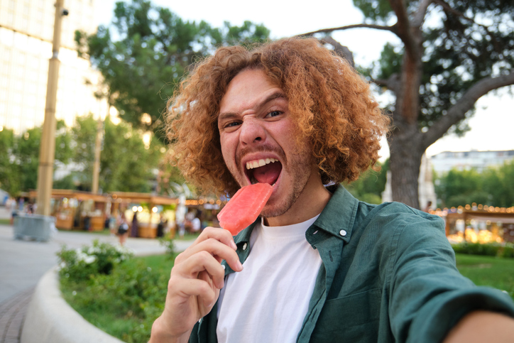 Young man with curly hair taking a selfie while biting a popsicle