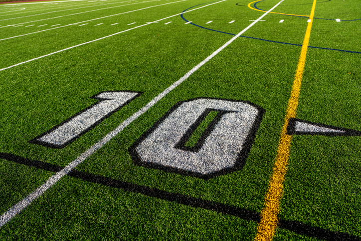 Green synthetic turf football 10 yard line, block number, in white along with black lacrosse line.