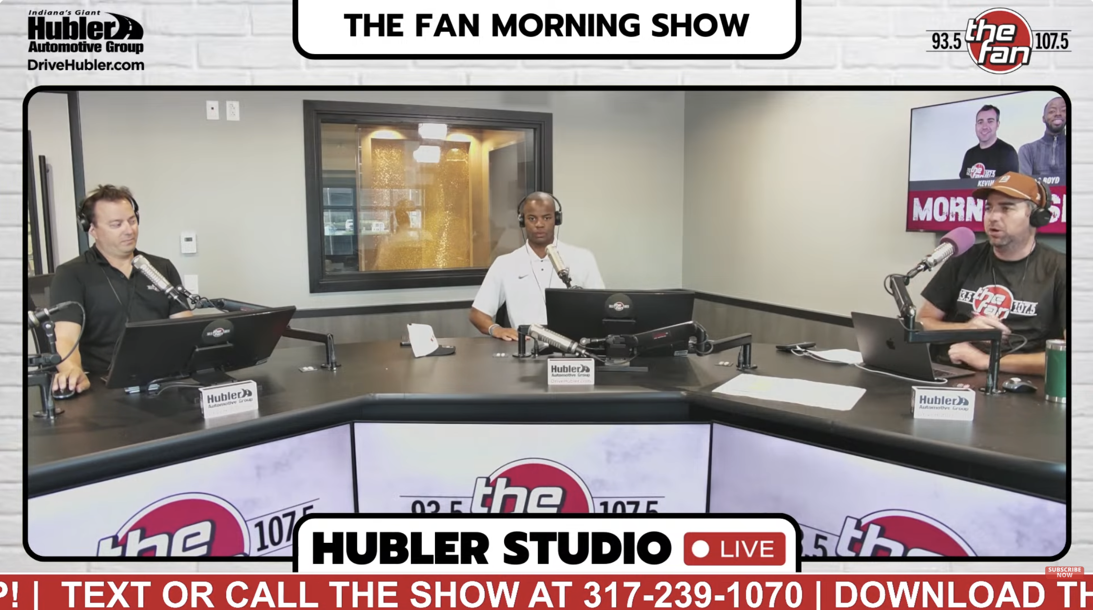 Three radio hosts seated at a studio desk with "The Fan Morning Show" branding on the wall behind them.
