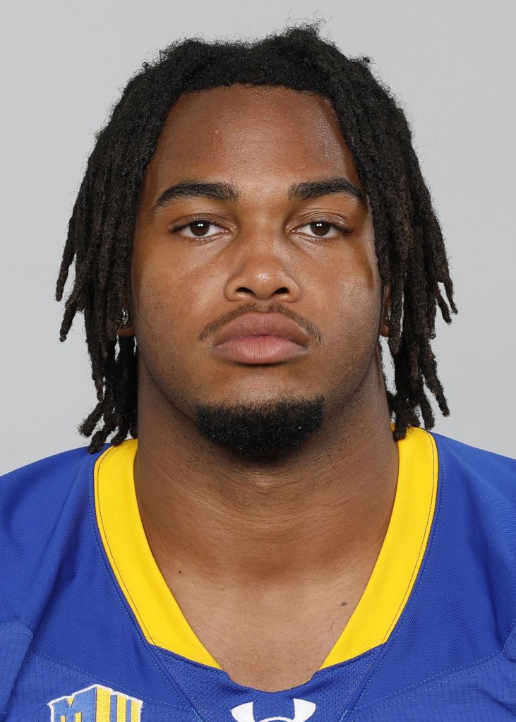 A close-up portrait of a young Black man with dreadlocks wearing a blue and yellow sports jersey.