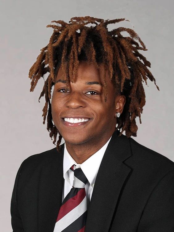 A smiling young man with dreadlocks wearing a black suit and tie.