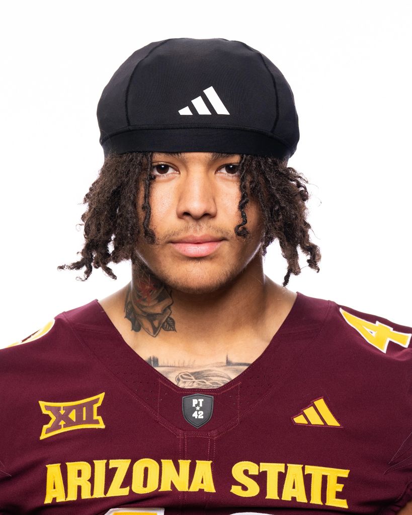 A person wearing a black Adidas cap and maroon Arizona State University jersey, with curly hair and tattoos.