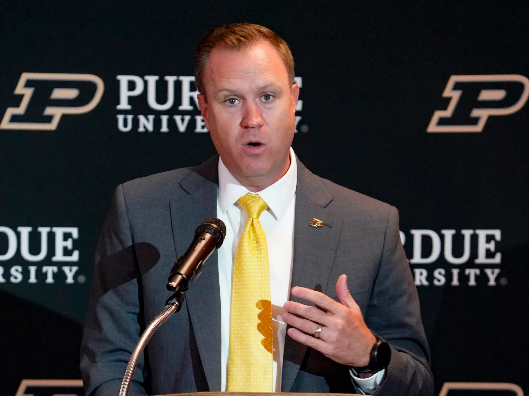 A man in a gray suit and yellow tie speaking at a podium with "Purdue University" branding visible behind him.