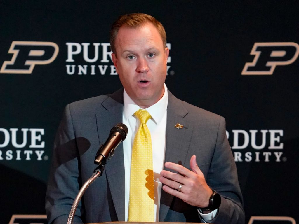A man in a gray suit and yellow tie speaking at a podium with "Purdue University" branding visible behind him.